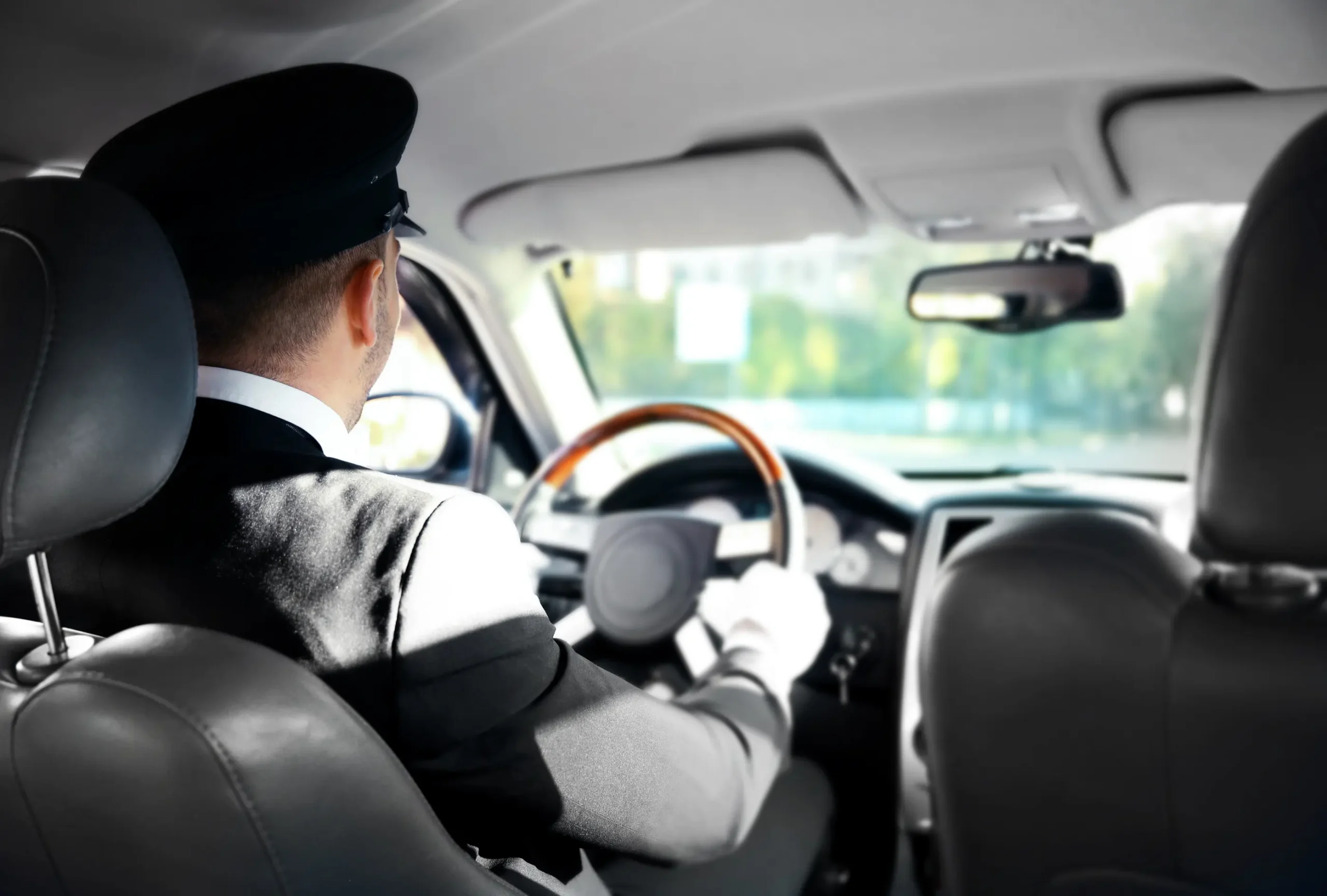 A chauffeur in a formal suit and cap drives a luxury car, gripping the steering wheel with gloved hands. Sunlight filters through the windshield.